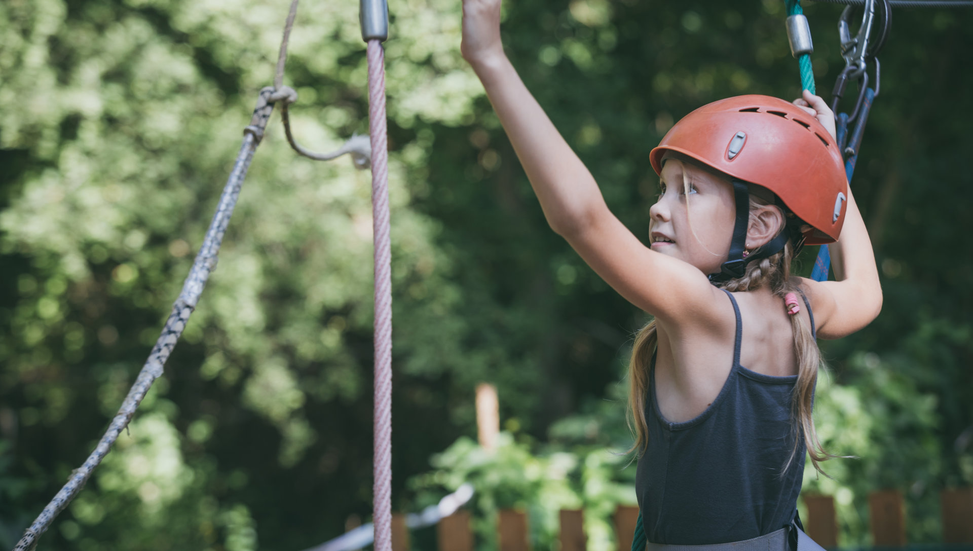 A young girl wearing a red helmet and harness climbs on a rope course in a forested area, looking focused and determined, with leafy trees in the background.