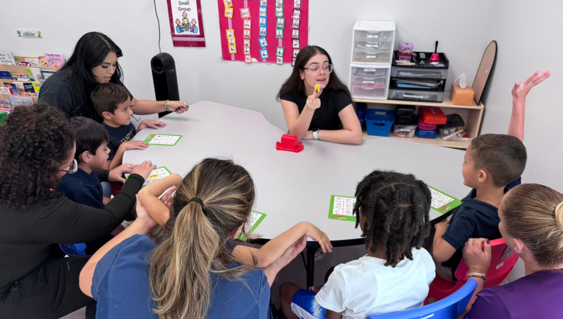 A group of children and two adults sit around a table in a classroom. 