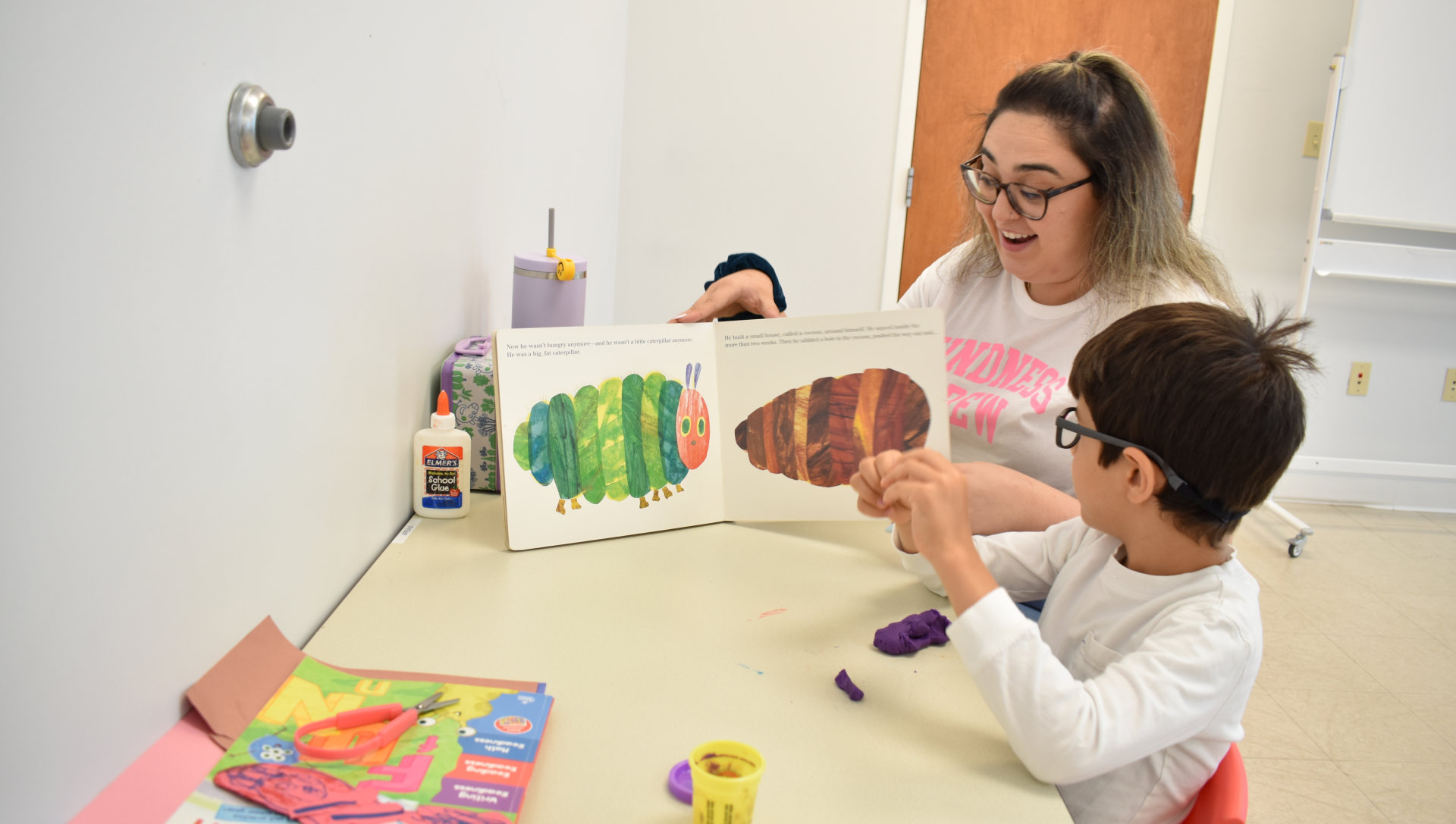 A woman reads a book to a child at a table with art supplies. 