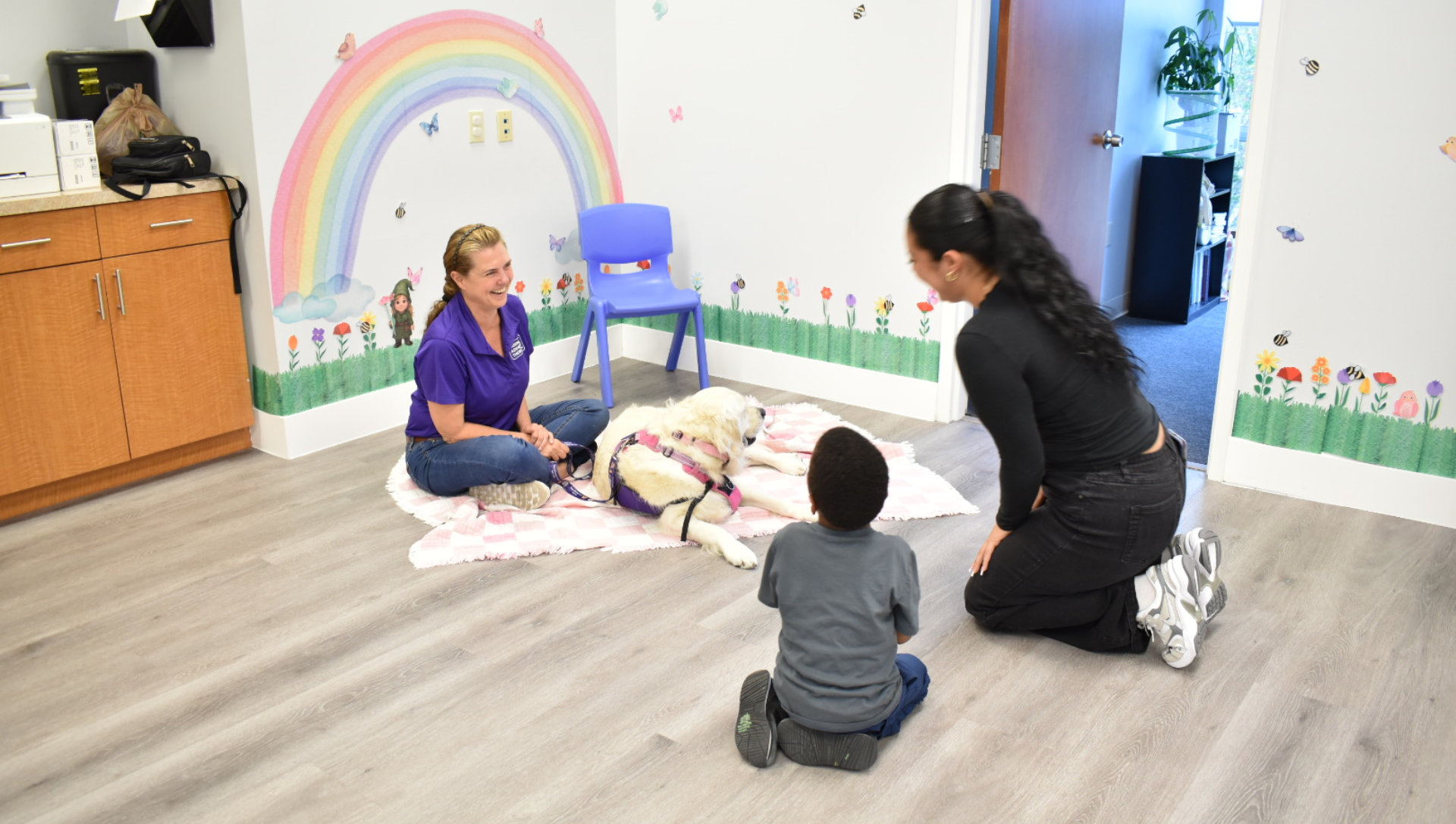 A therapy dog lays on a blanket in a colorful room with a rainbow mural. A woman in a blue shirt sits with the dog, while another woman and a child kneel nearby.